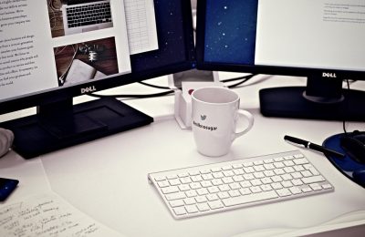 工作台上并排摆放的三台显示器，屏幕里是不同角度的3d模型线框与贴图预览，暖色台灯照亮键盘，氛围专注安静